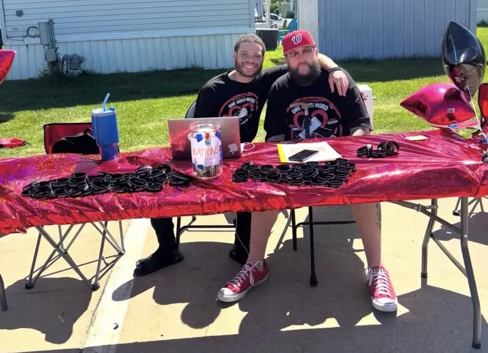 Cody Greggs-Dorsey and Jason Lewis in One Good Word One Good Deed shirts behind an outdoor table covered with wristbands.