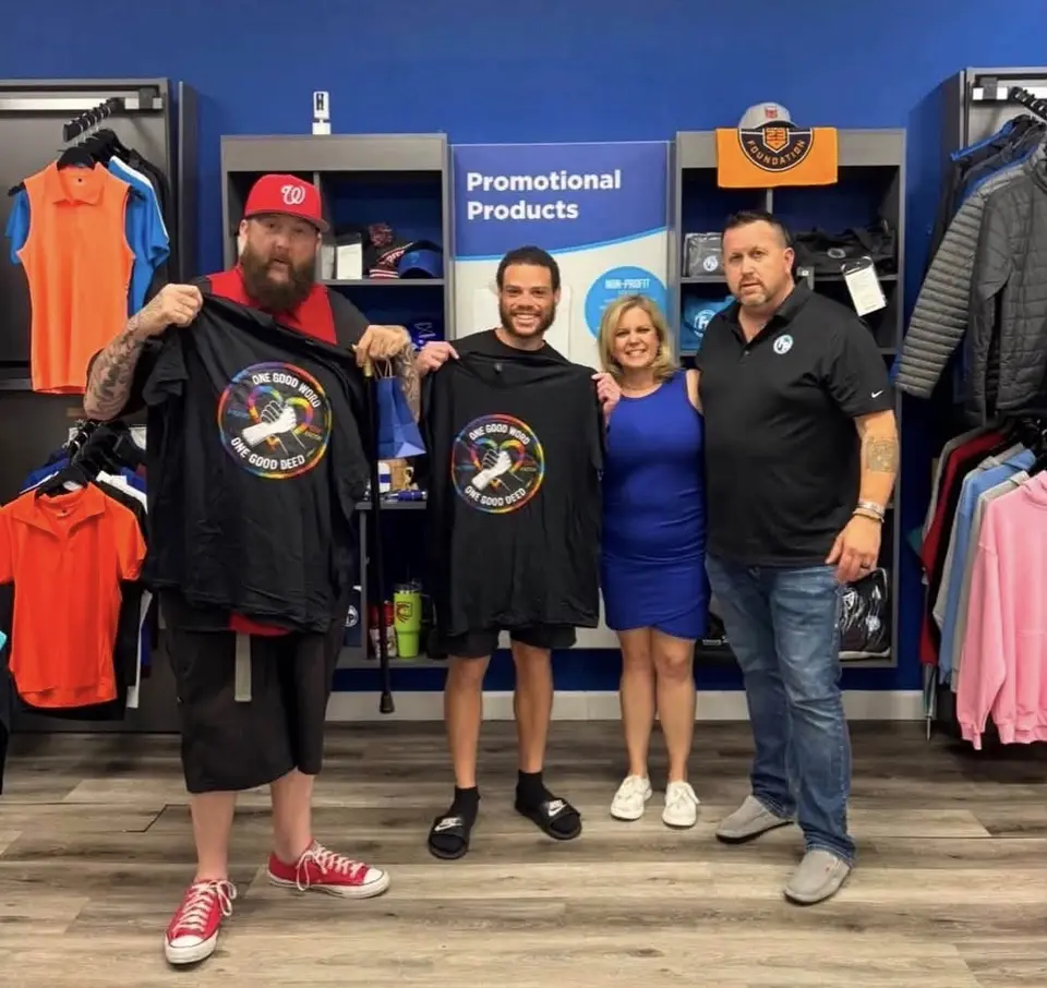 Jason Lewis and Cody Greggs-Dorsey holding up One Good Word One Good Deed shirts with two other people in a promotional products store.